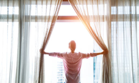 Мужчина открывает шторы в светлой комнате, обращенной к солнечному свету, что символизирует новые начинания и позитивный настрой.