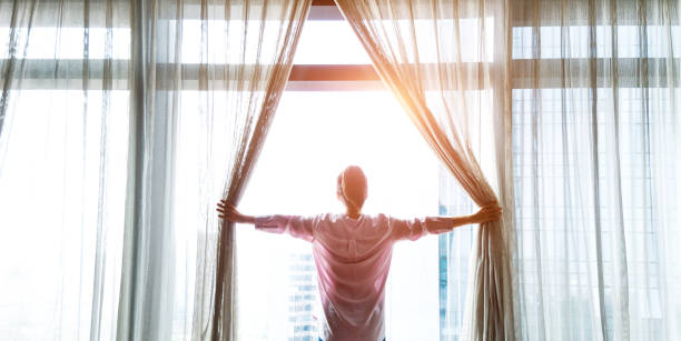 Мужчина открывает шторы в светлой комнате, обращенной к солнечному свету, что символизирует новые начинания и позитивный настрой.