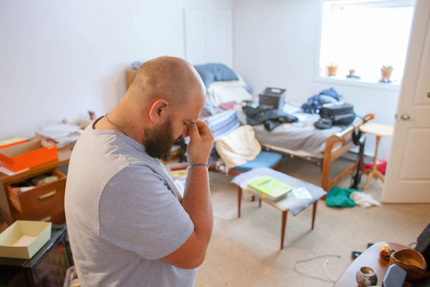 Лысый мужчина в серой рубашке выглядит напряженным в грязной спальне с разбросанными вещами и открытыми ящиками.