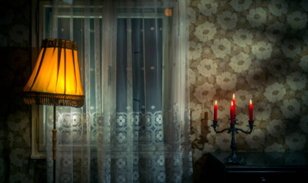 Винтажный интерьер комнаты с зажженной настольной лампой и горящими свечами на комоде, отбрасывающими теплое сияние на цветочные обои и прозрачные шторы