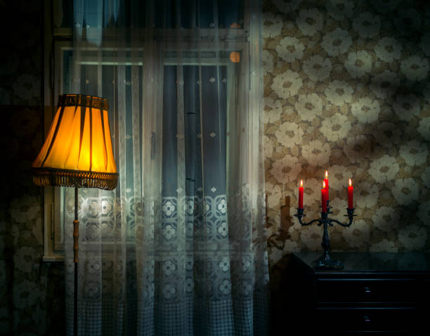 Винтажный интерьер комнаты с зажженной настольной лампой и горящими свечами на комоде, отбрасывающими теплое сияние на цветочные обои и прозрачные шторы